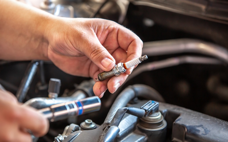 A person standing in front of a vehicle with its hood open. The person holds a small spark plug in their left hand.