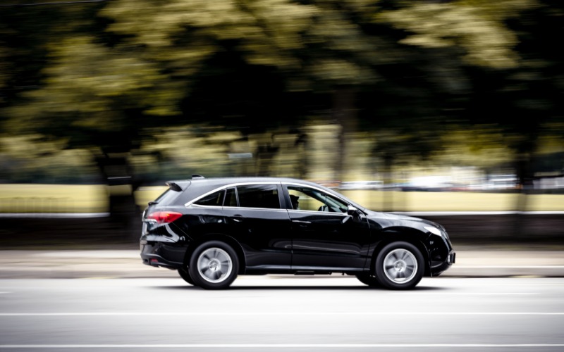 A black car driving down the road during the daytime. The car drives fast past many trees and an open field.