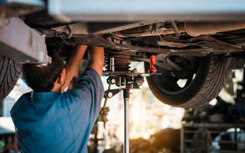 A mechanic working on the underside of a large car that is raised high in an automotive repair shop.