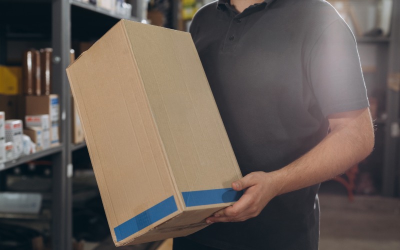 An employee holding a cardboard package in a large commercial storage space. The box has a blue strip at the bottom.
