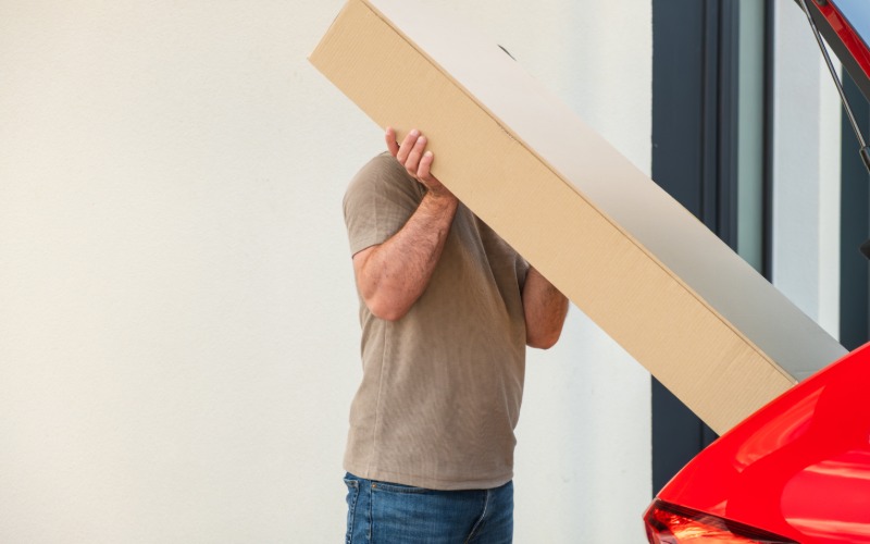 A person wearing a brown shirt removes a long cardboard package from the trunk of their bright red car.