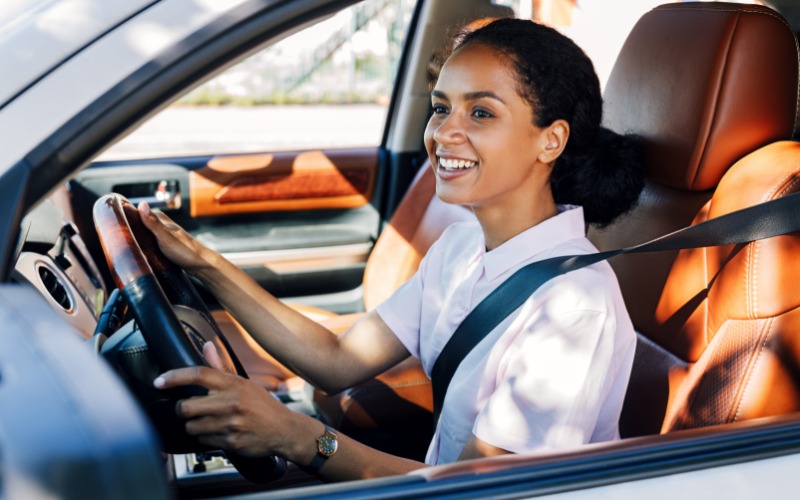 A person wearing a white shirt smiling and driving a car with the driver-side window down on a sunny day.