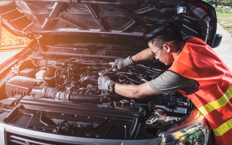 A mechanic wearing a high-visibility vest using a wrench to make repairs under the hood of a car parked outside.