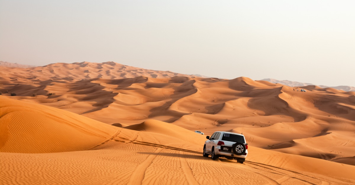  A white car driving through the desert sands on a sunny day. Two more white cars wait in the distance.