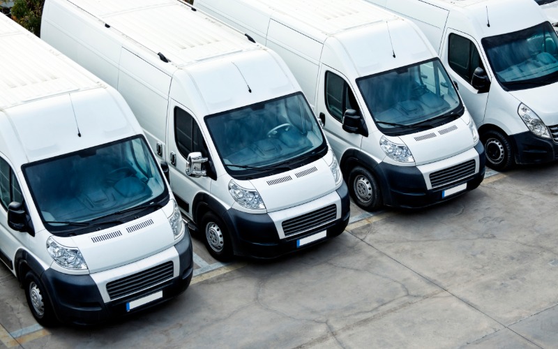 A fleet of five identical white, box-shaped commercial vans parked outside in a line in a parking lot.