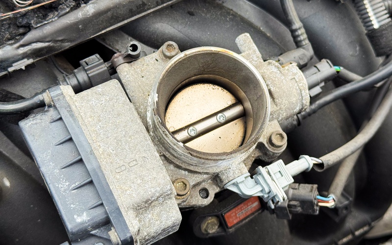 A car engine throttle body with many automotive connectors attached to it. The throttle body is gray and worn.