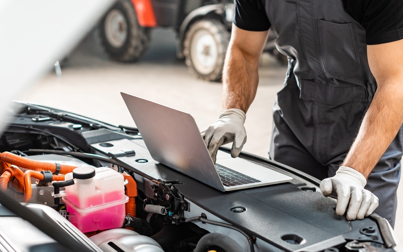 A person wearing gray gloves while using a laptop in front of a car. The car has its hood propped open.