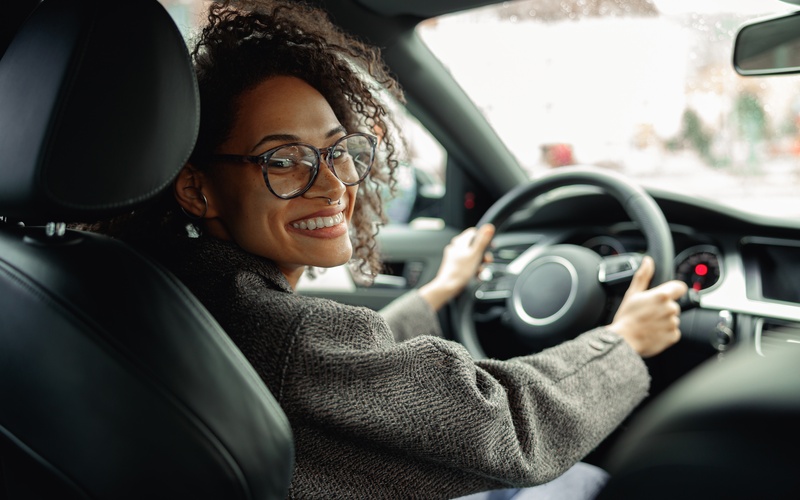 A person wearing glasses is gripping the steering wheel of a car and smiling while looking to the right.