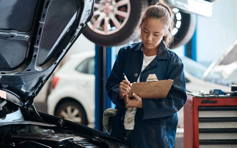 A mechanic in blue coveralls and a white shirt writing on a clipboard with a white pen in an automotive garage.